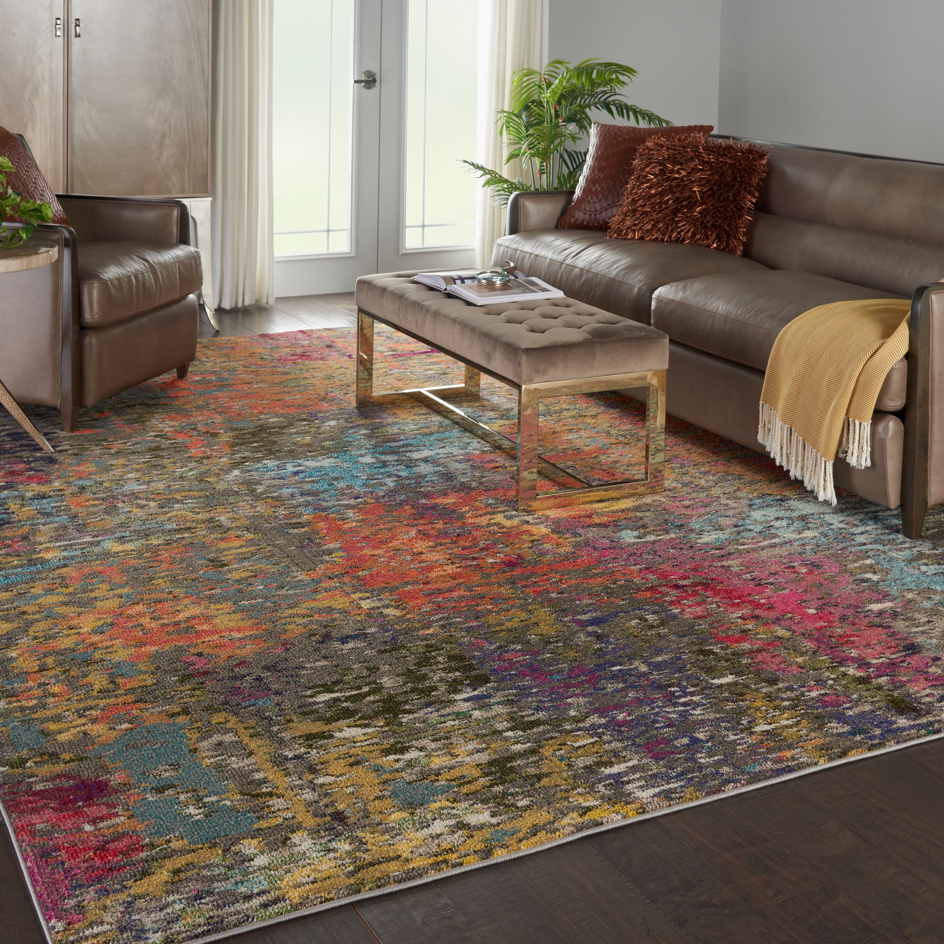 Living room with a vibrant multicolor abstract rug, taupe tufted ottoman on gold legs, and brown leather sofa.