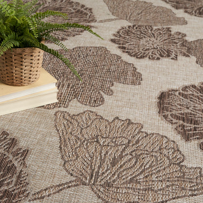 Beige rug with large brown botanical leaf pattern; woven texture; small potted fern in wicker basket atop a stack of books.