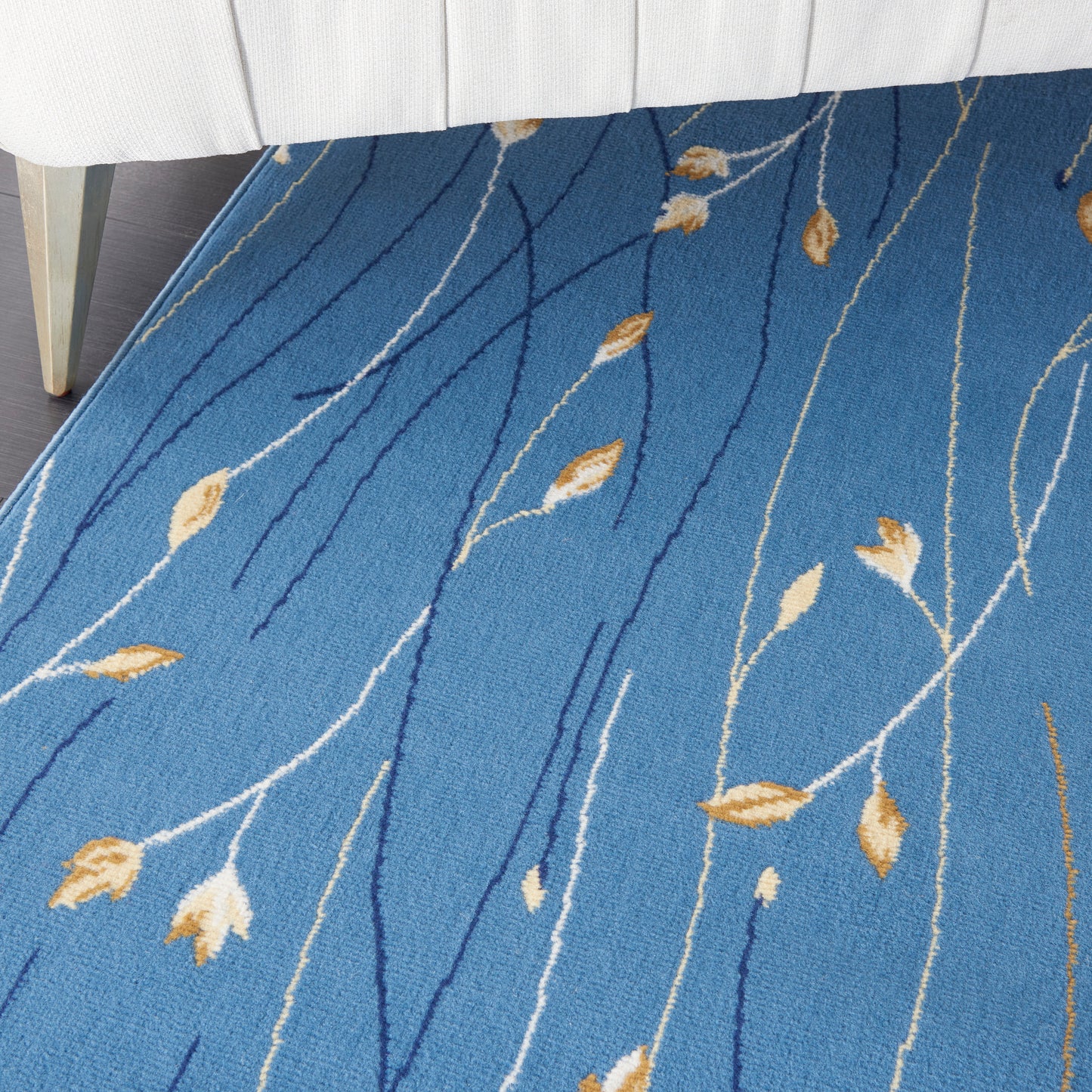 Blue area rug with white and dark blue wavy stems and tan leaf motifs under a white upholstered bed.