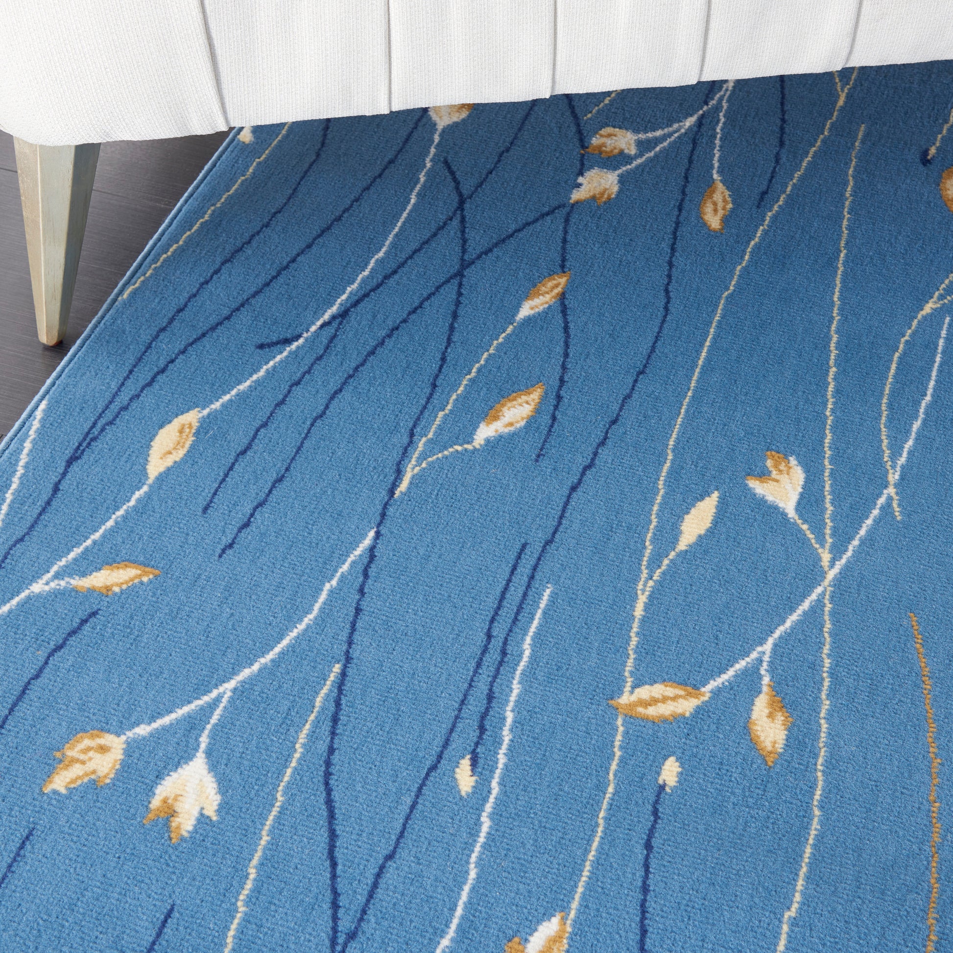 Blue area rug with white and dark blue wavy stems and tan leaf motifs under a white upholstered bed.