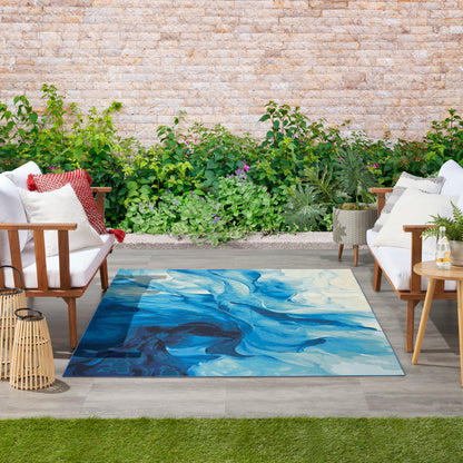 Outdoor patio rug with blue abstract wave pattern, flanked by white cushioned wooden armchairs amid lush greenery.