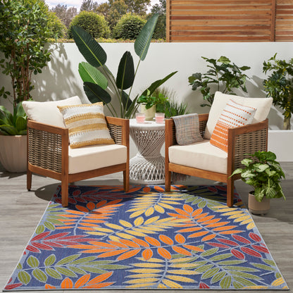 Outdoor patio rug with tropical leaf pattern; visible are wooden armchairs with cream cushions, striped pillows, and a round white metal table.