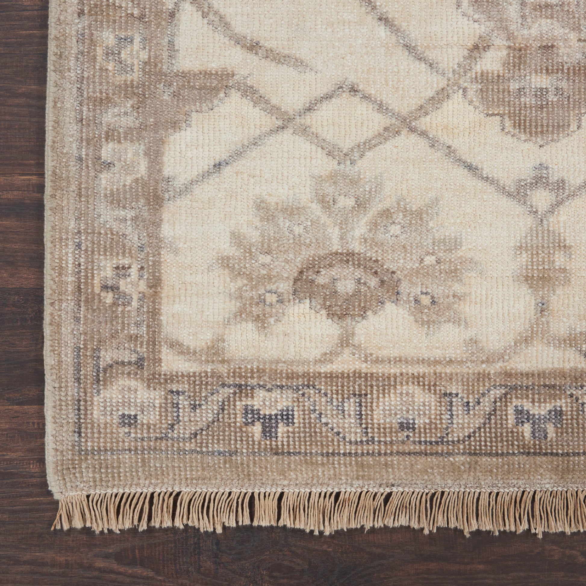 Beige oriental-style rug with floral border and fringed edge on dark wood floor.