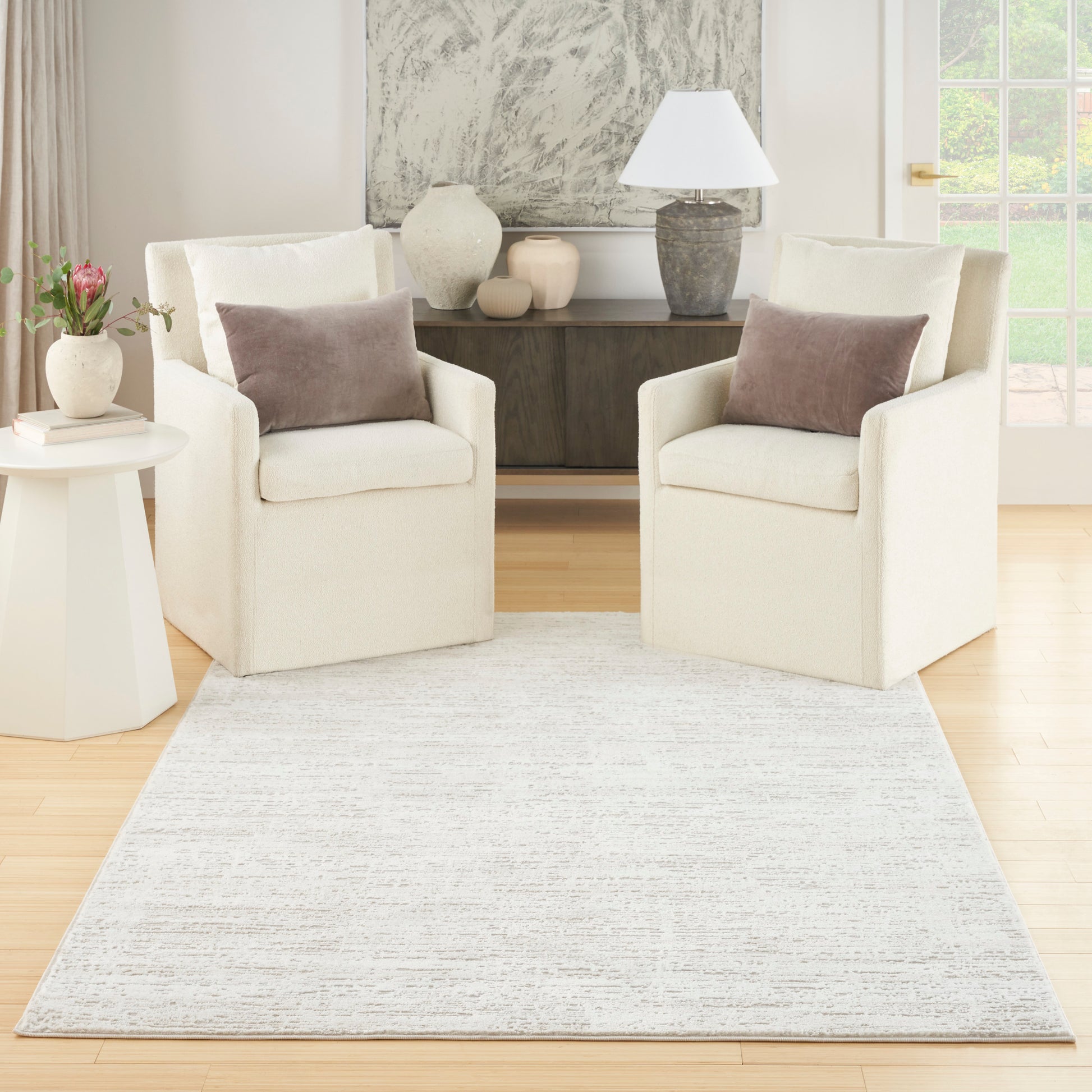 Two cream fabric armchairs with taupe cushions sit on a light textured rug in a neutral living room with a wooden console.