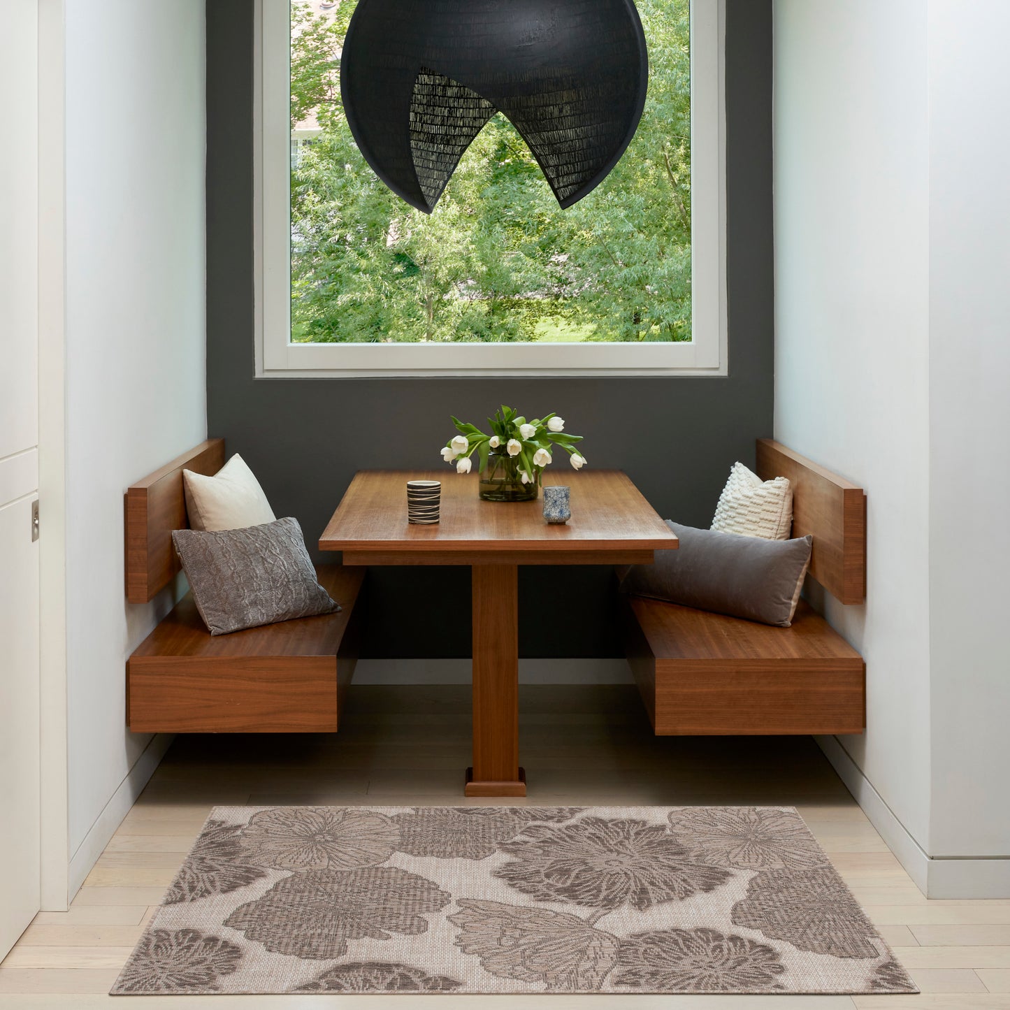 Tropical rug in a wood dining nook with benches and a square pedestal table, grey walls, a black pendant lamp, and a vase of flowers.