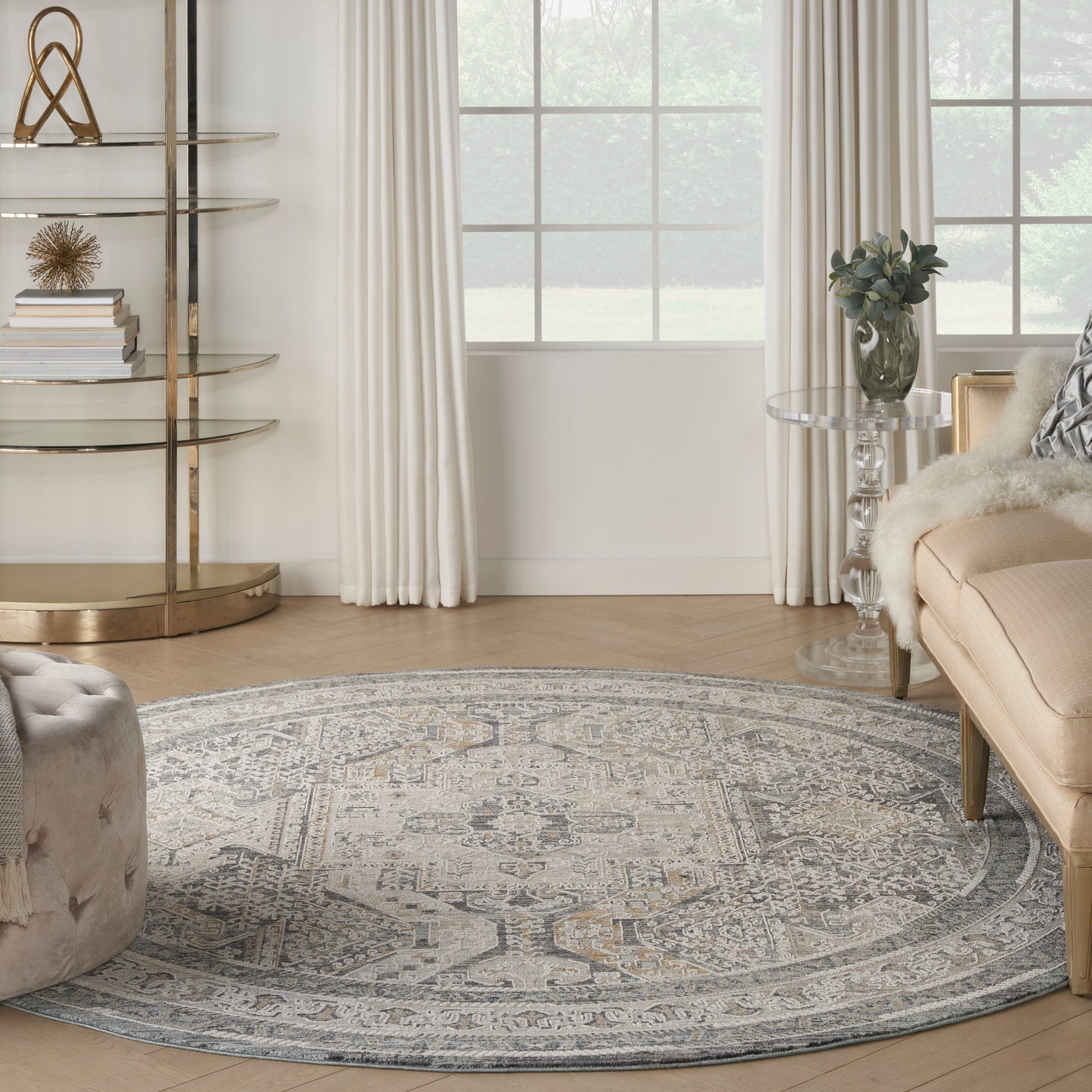 Round, muted gray-beige area rug in a light living room with beige sofa, glass side table, and gold metal shelves.