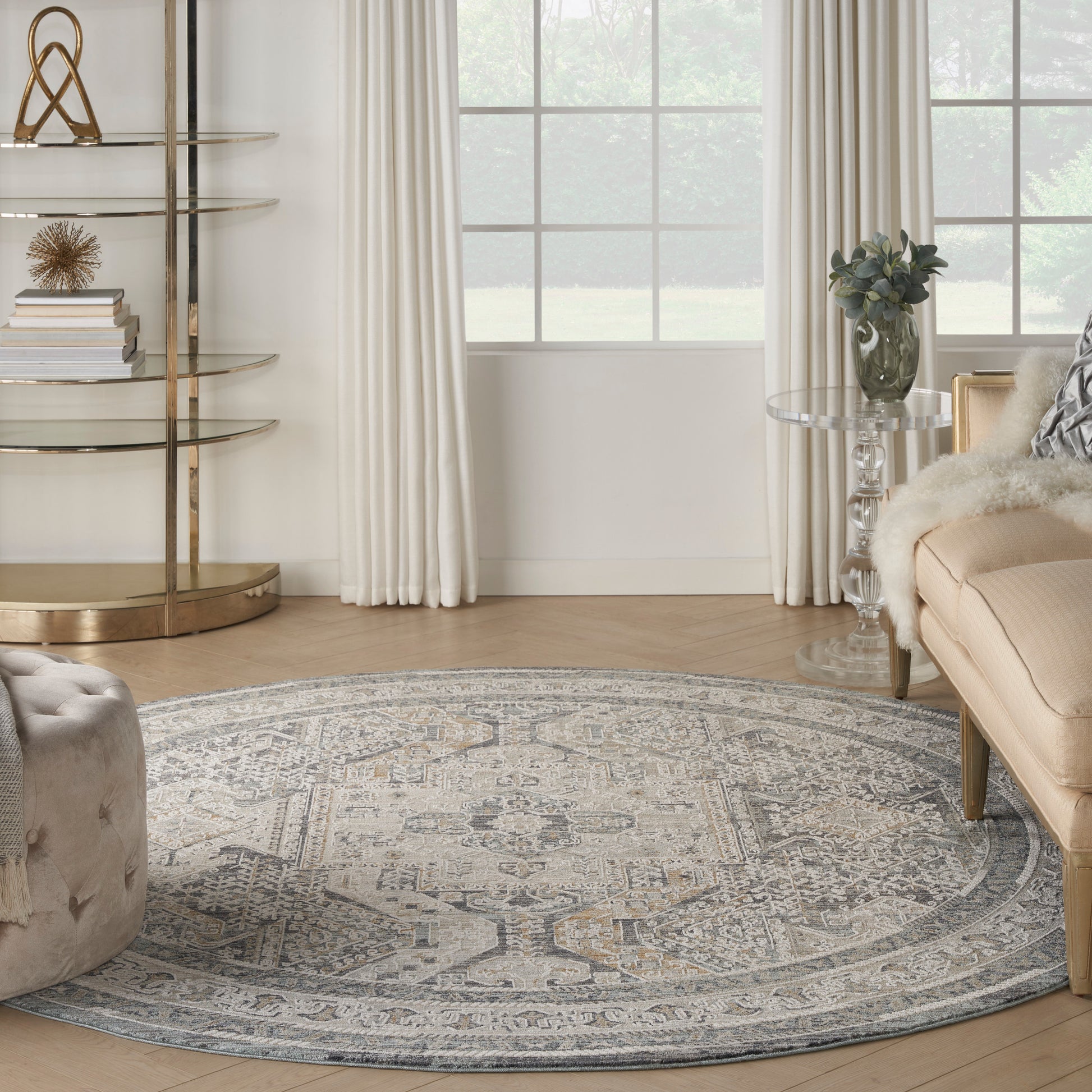 Round, muted gray-beige area rug in a light living room with beige sofa, glass side table, and gold metal shelves.