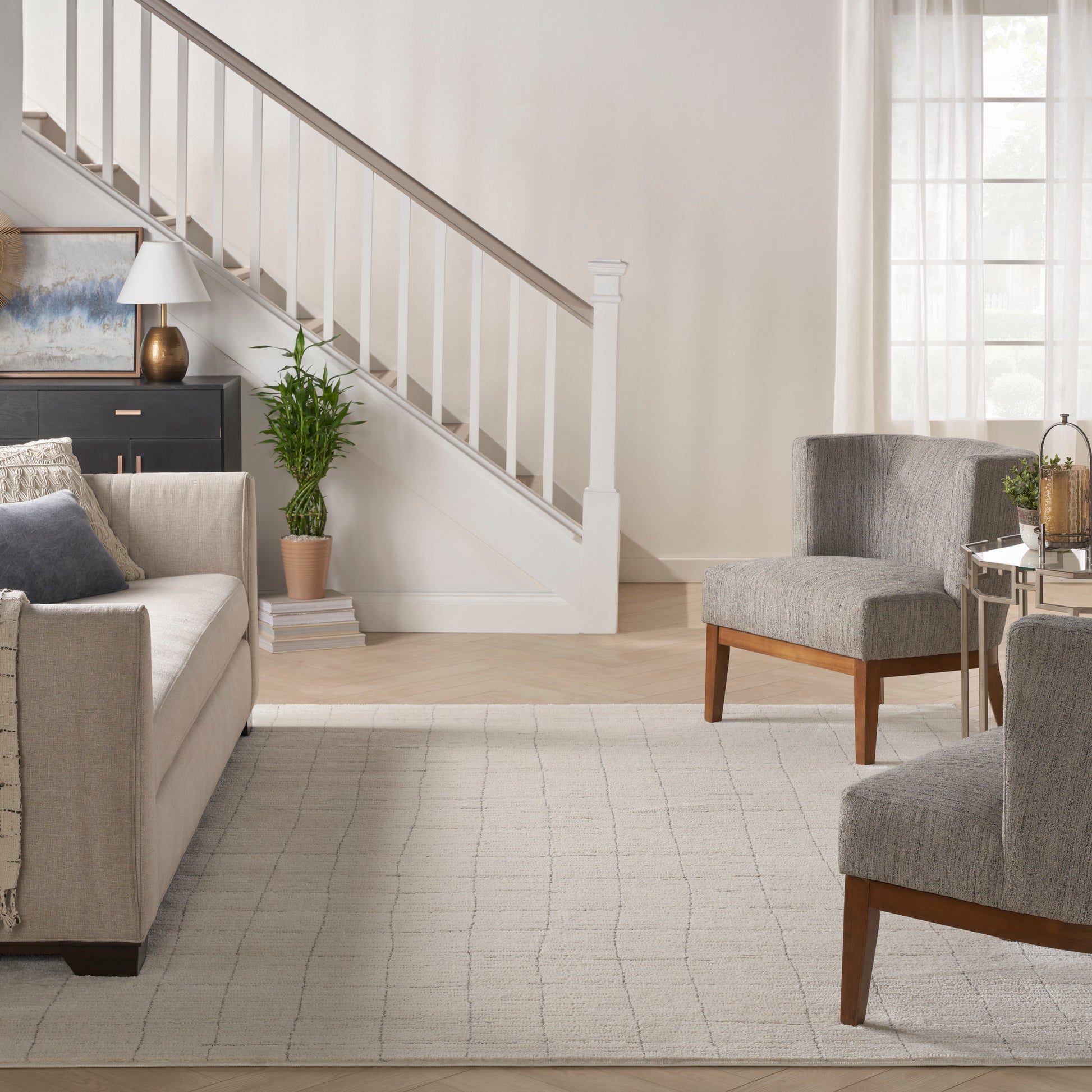 Bright living room with staircase, beige sofa, gray armchairs, light rug, potted plant, glass coffee table, sheer curtains.
