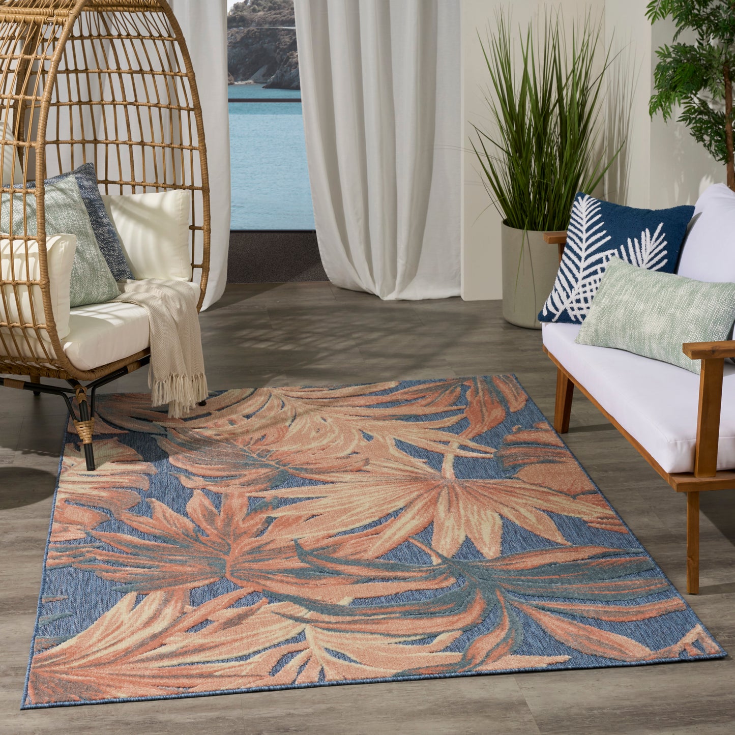 Coastal living room with a rattan egg chair, white sofa, tropical blue-peach leaf rug, & potted plants by sheer curtains.