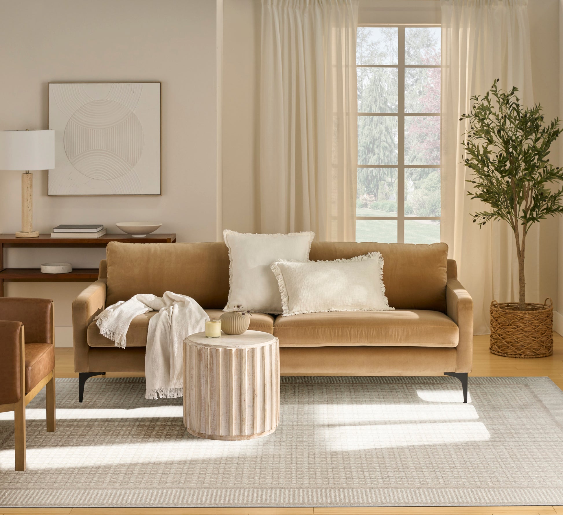 Beige rug in a living room scene with a tan leather sofa and white pillows, cream throw; round slatted wood coffee table; potted tree in a wicker basket.