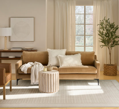 Beige rug in a living room scene with a tan leather sofa and white pillows, cream throw; round slatted wood coffee table; potted tree in a wicker basket.