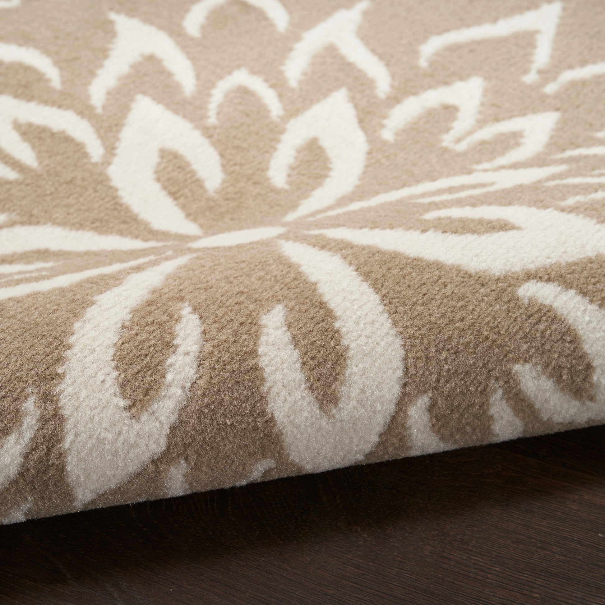 Beige plush rug with white floral motif and bold medallion pattern, folded edge on dark wood floor.