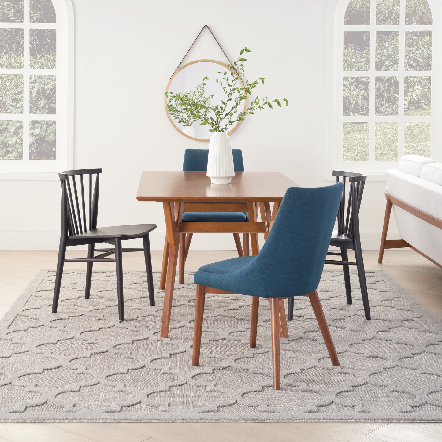 Mid-century wooden dining table with blue upholstered chairs and black wood chairs, white vase with greenery, round mirror, gray patterned rug.