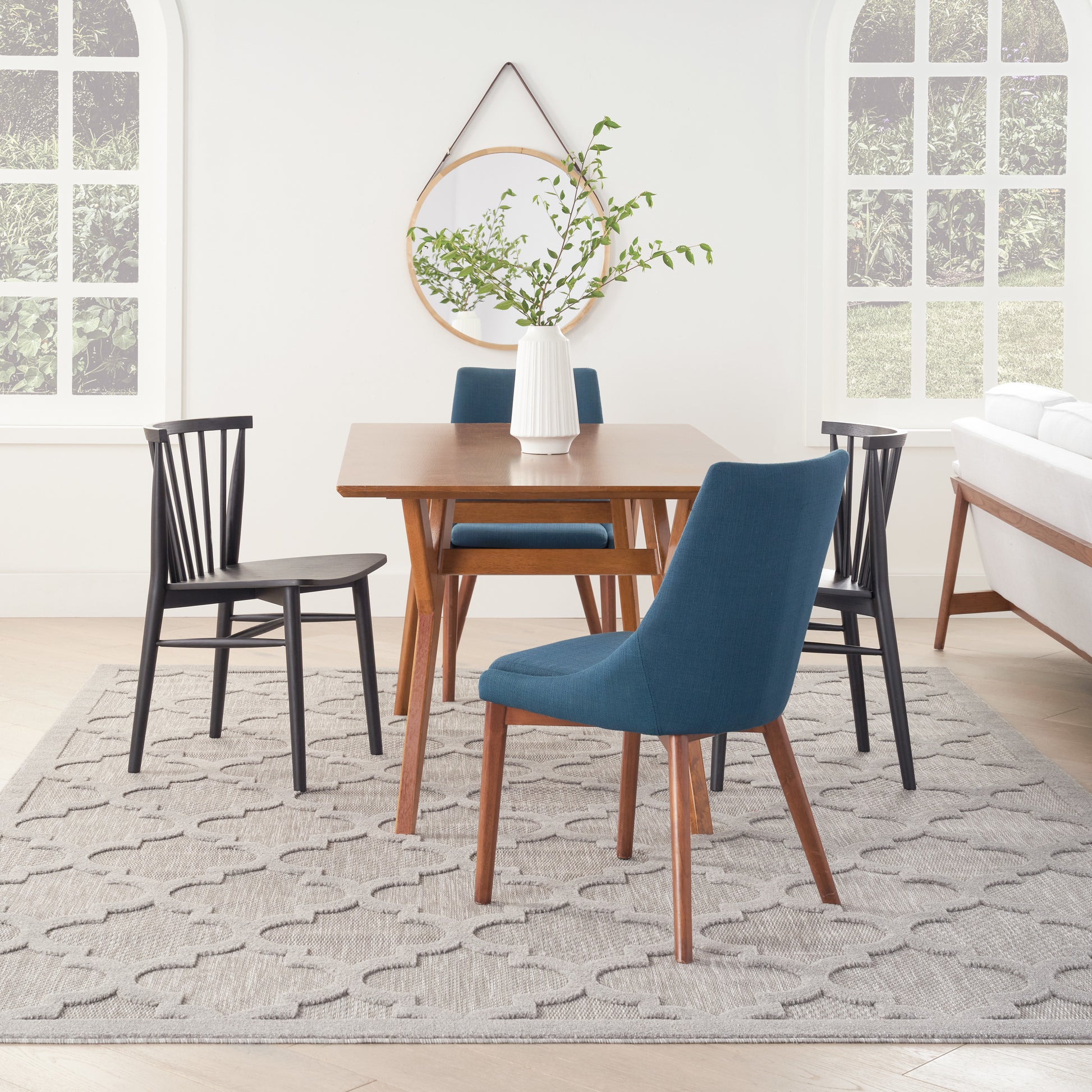 Mid-century wooden dining table with blue upholstered chairs and black wood chairs, white vase with greenery, round mirror, gray patterned rug.