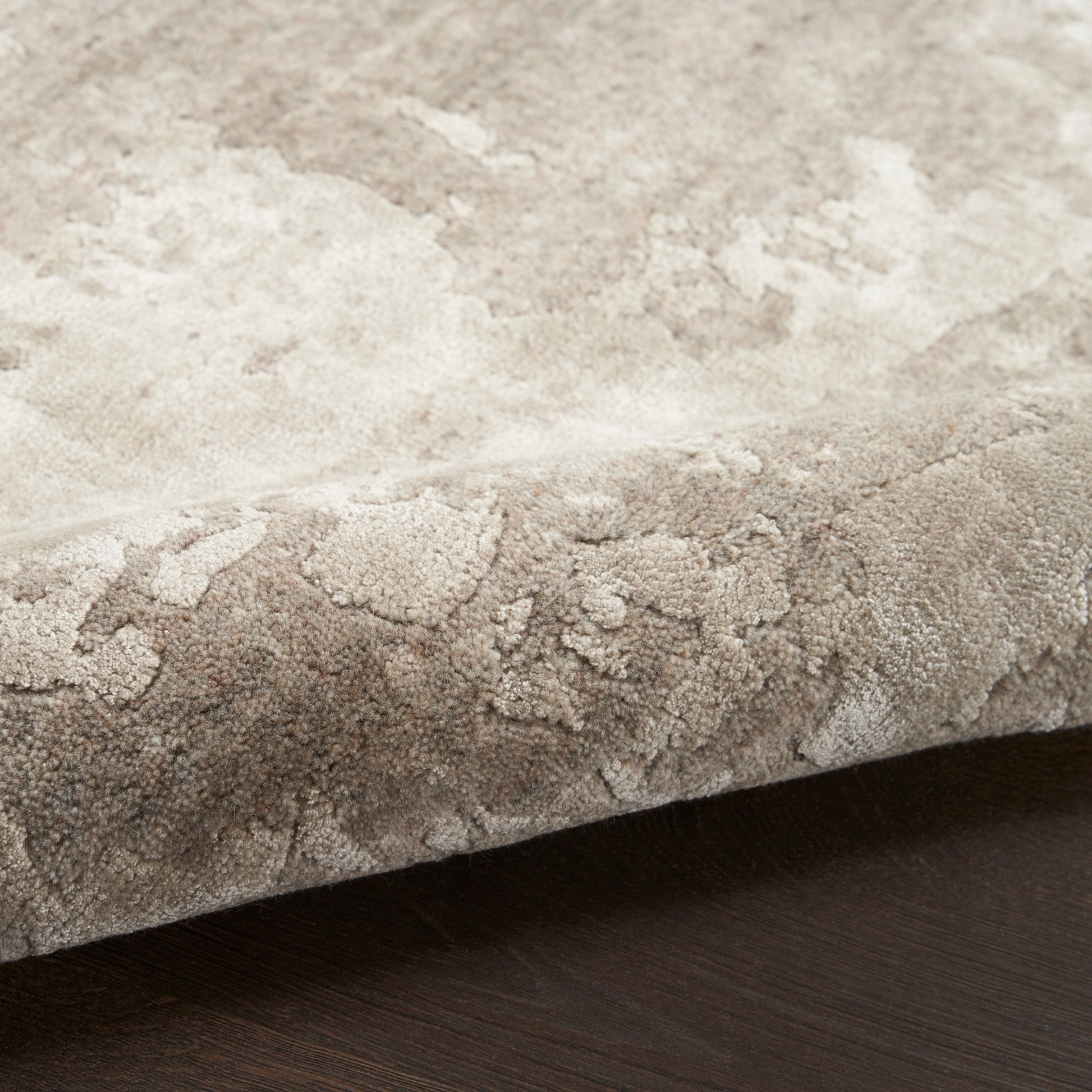 Soft plush beige rug with subtle mottled pattern, edge curled on dark wood floor.