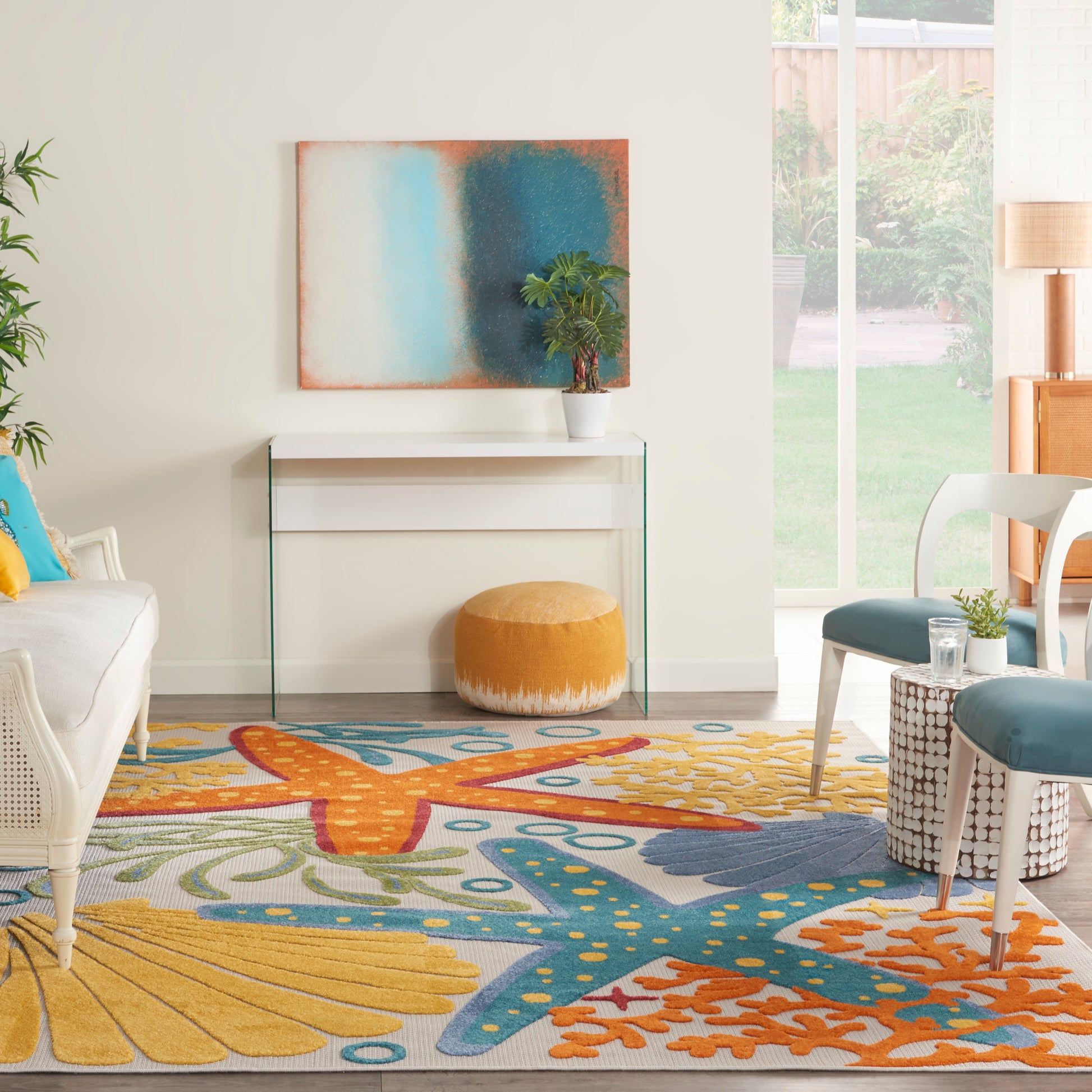 Bright living room with a sea-themed rug in yellow, blue, and orange; glass console, mustard pouf, wicker chair, and plant.
