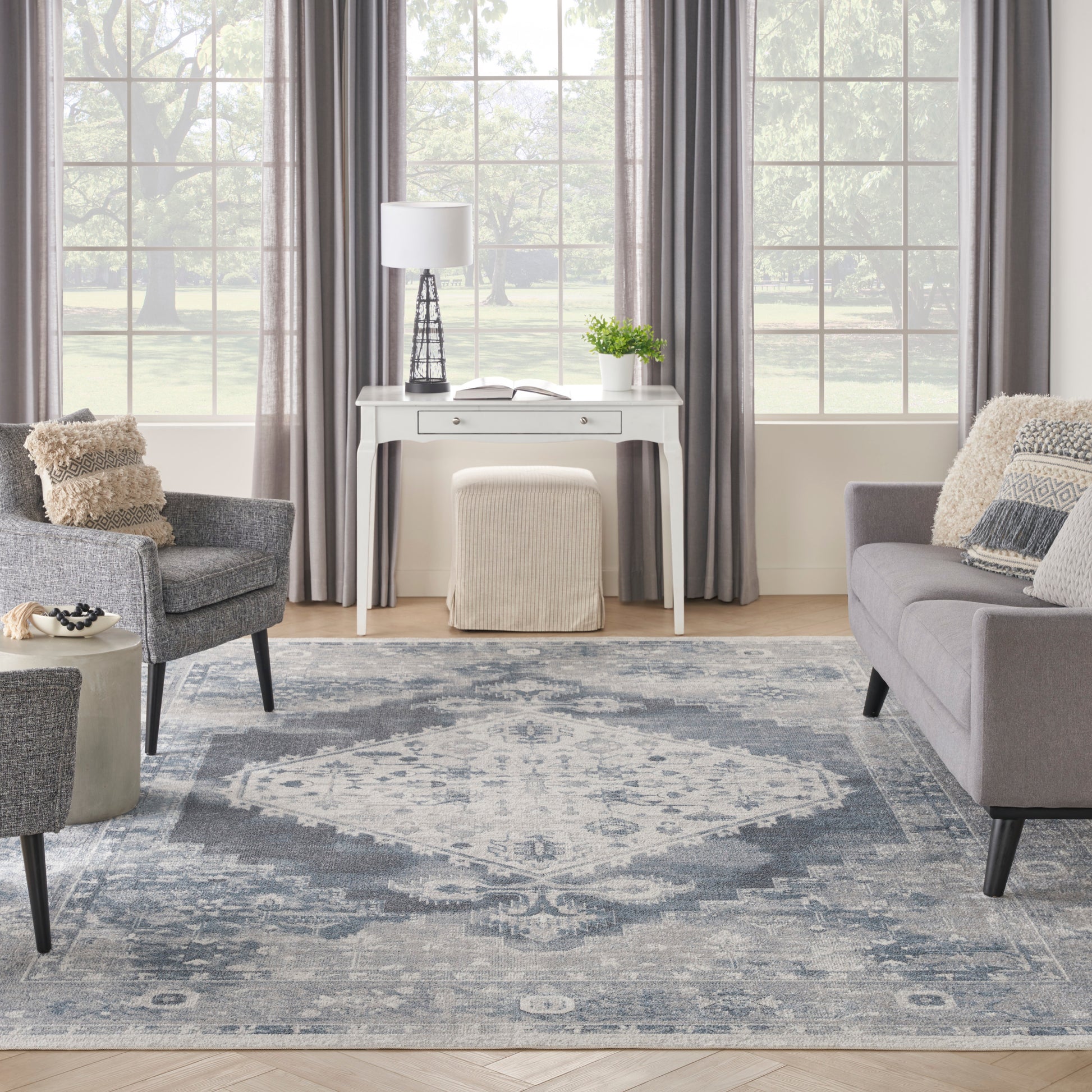 Cozy gray living room with a blue patterned area rug, white console table, gray sofa and armchairs, cream pouf, lamp.