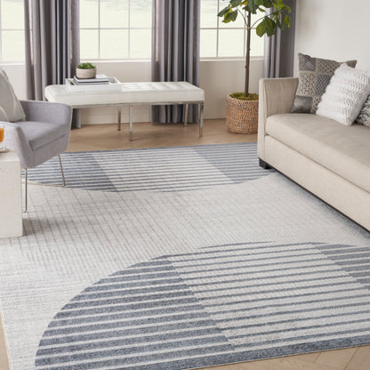 Blue-gray geometric area rug with curved stripes, in a modern beige living room with sofa, chairs, and a potted plant.
