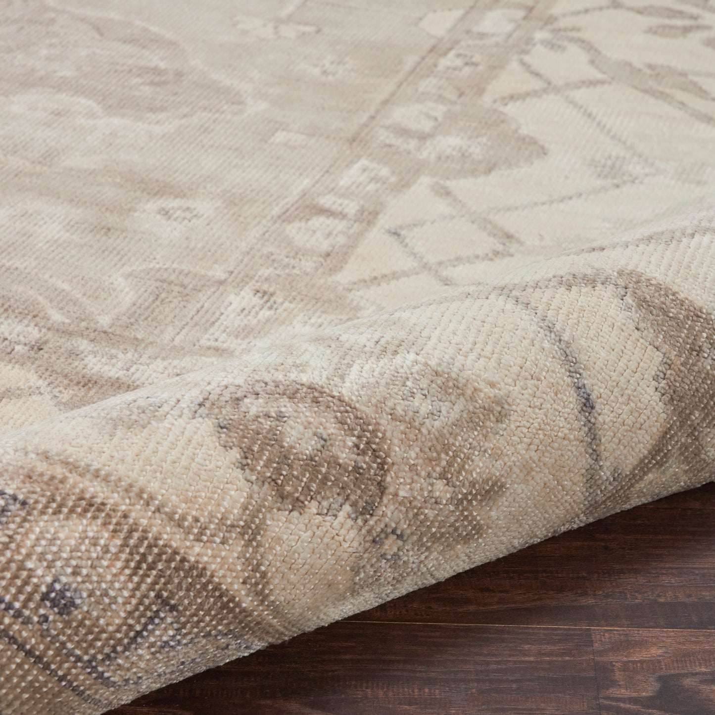 Beige woven rug with muted geometric/oriental pattern, textured fabric, draped over dark wood floor.