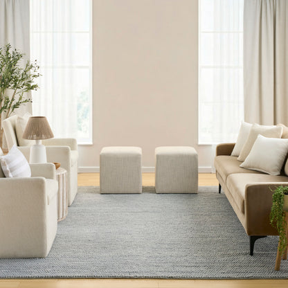 Neutral living room with cream fabric sofas, two beige ottomans, gray rug, plant, and soft natural light.