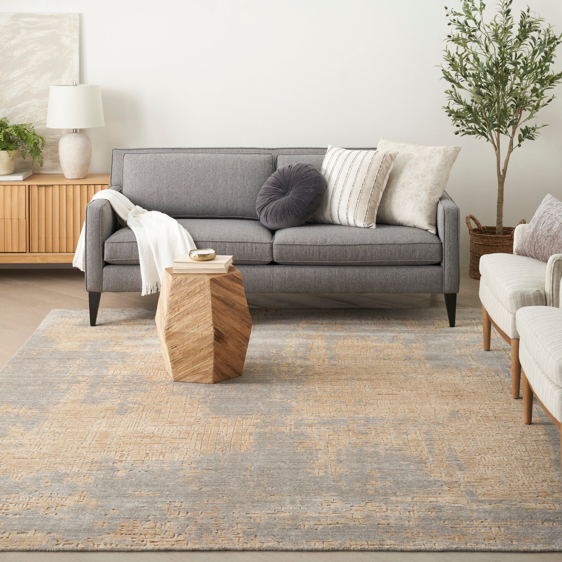 Modern gray fabric sofa with neutral cushions and white throw, geometric wooden coffee table, beige-blue rug, plant.