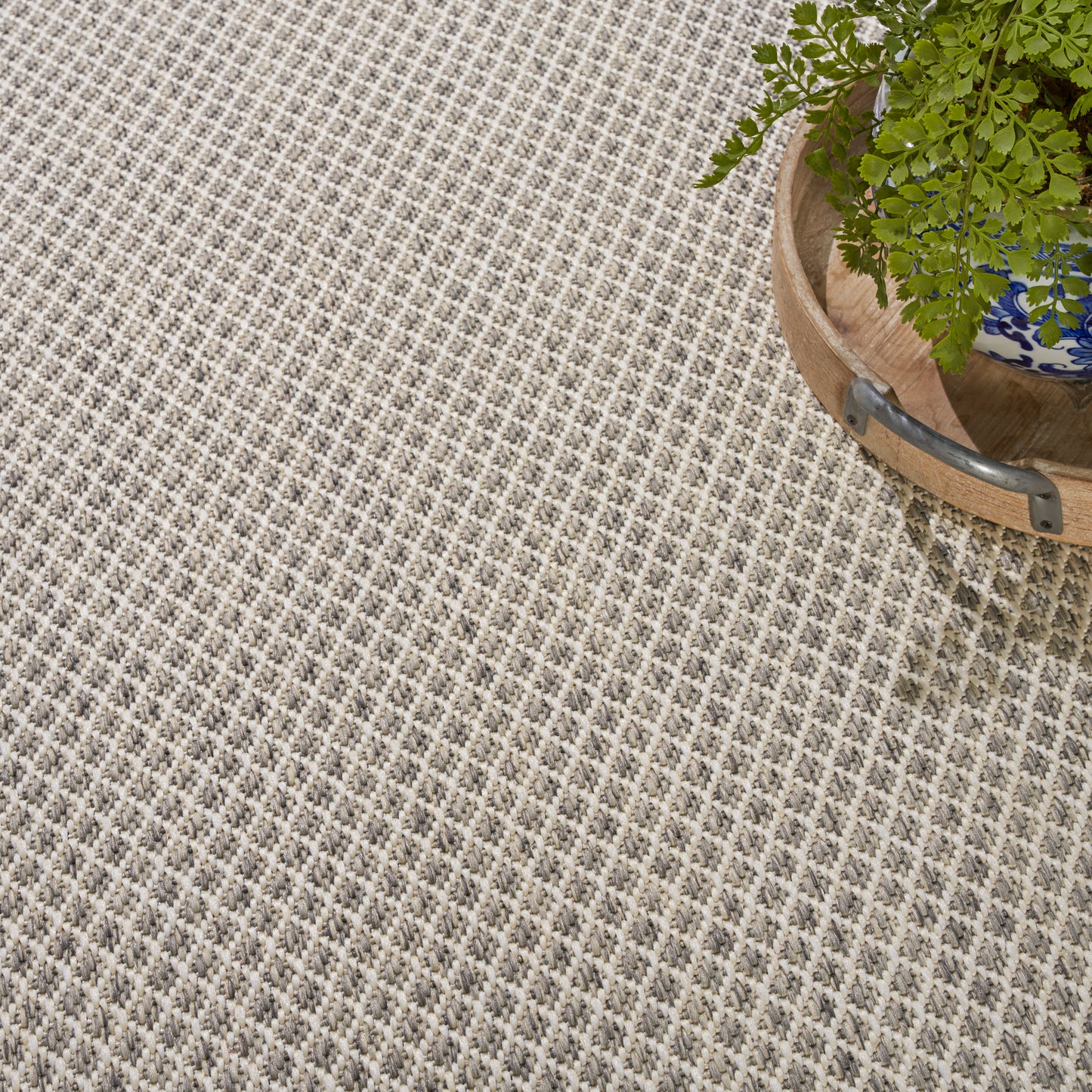 Beige textured rug with small repeating geometric pattern; wooden barrel planter with a green fern in the top-right.