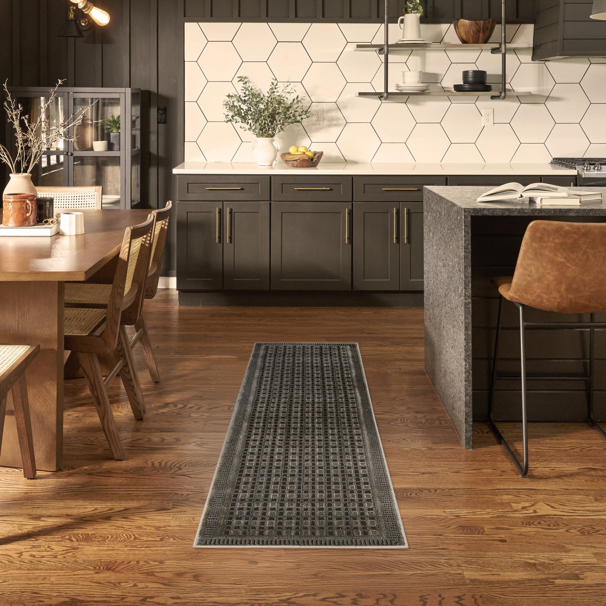 Long dark patterned runner rug on hardwood floor in a modern kitchen with black cabinets, white hex tile backsplash.