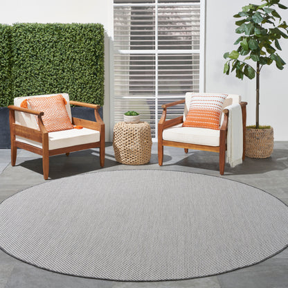 Outdoor patio: two wooden armchairs with white cushions, orange pillows, round gray rug, wicker side table.