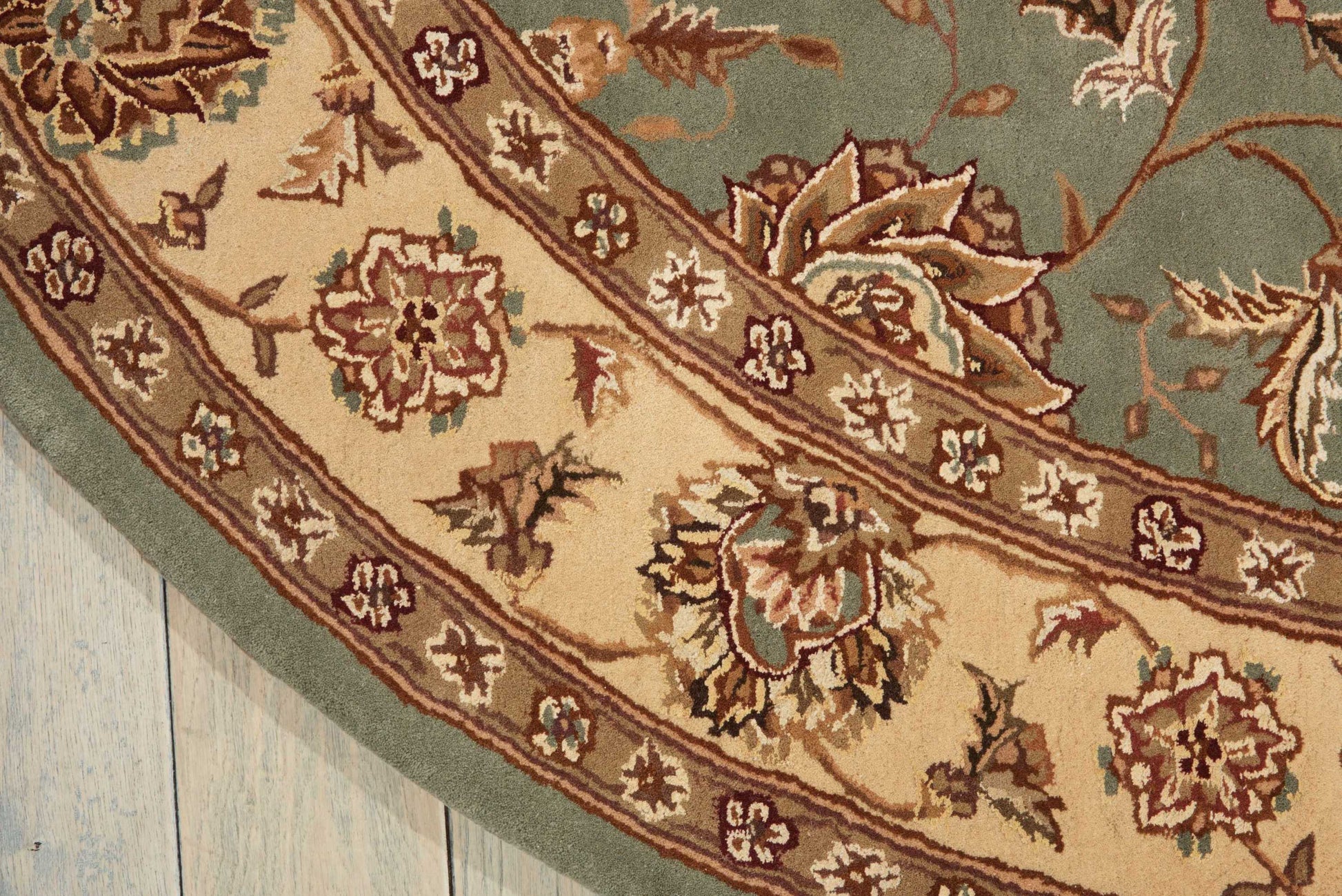 Partial view of a woven oriental rug: teal field, beige border with floral motifs, curved edge on light wood floor.