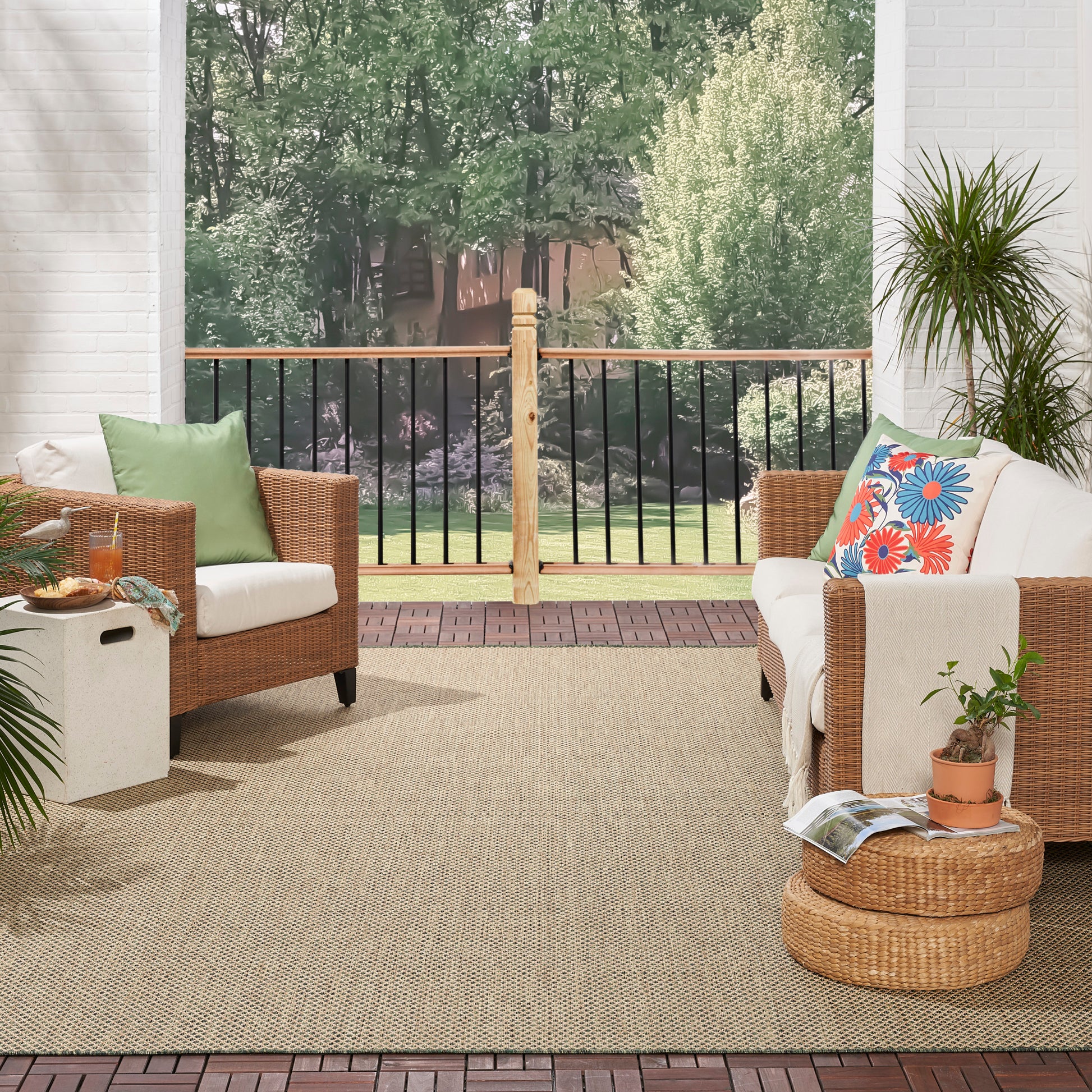 Outdoor wicker seating on a beige rug with white cushions, green and floral pillows, round ottomans and potted plants.