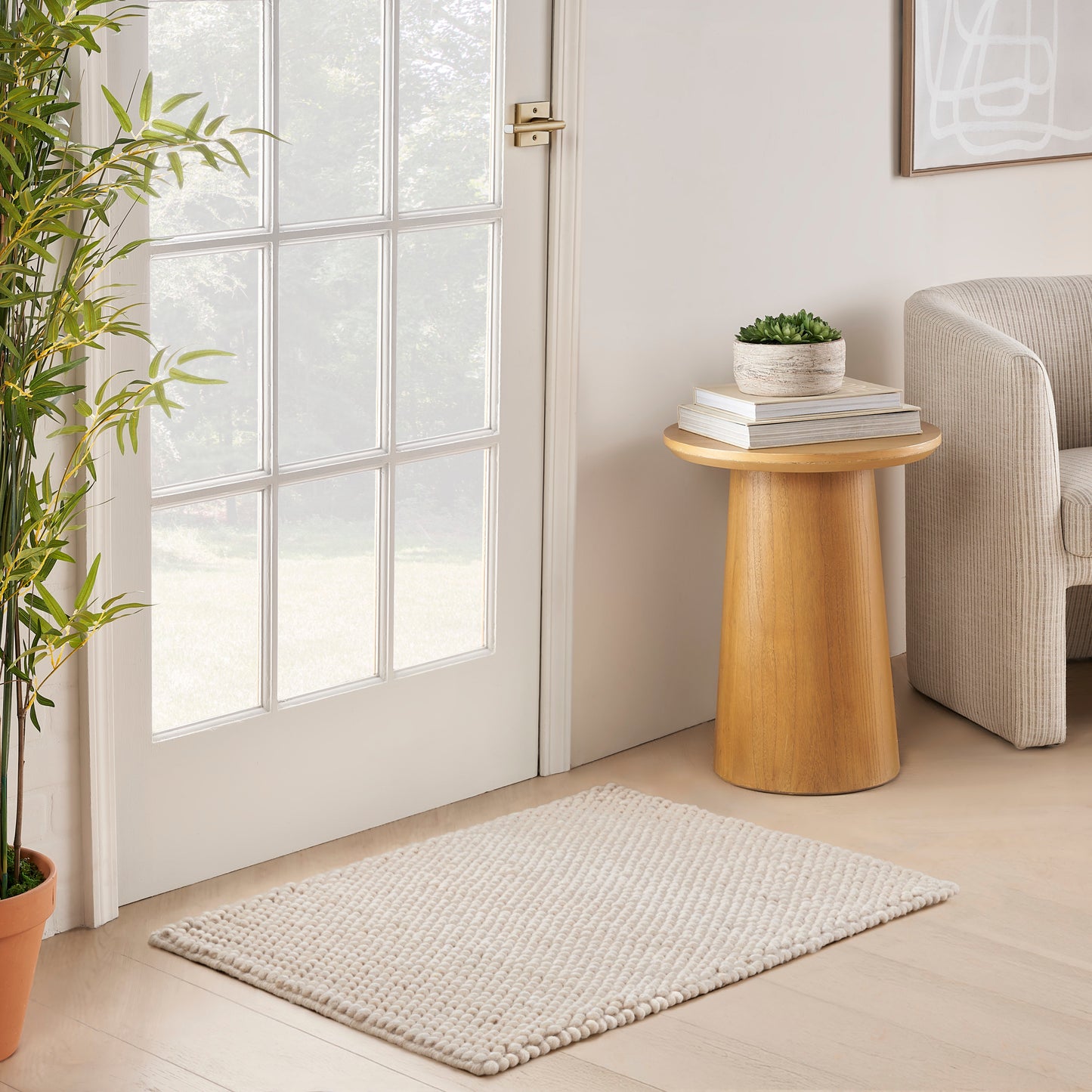 Round wooden side table with books and a small plant beside a beige sofa; bamboo plant by glass-pane door and cream rug.