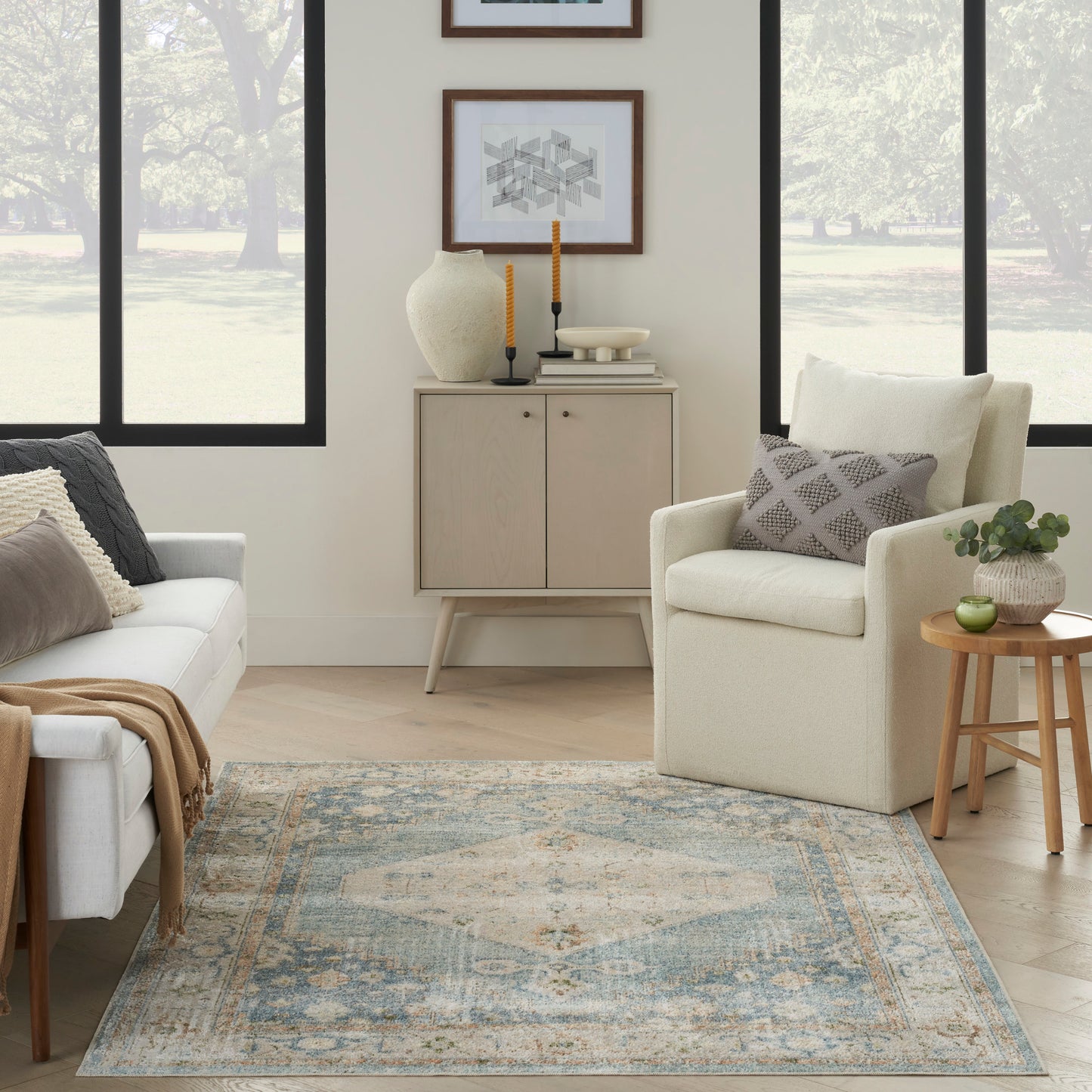 Neutral living room with beige armchair, light wood cabinet, blue distressed rug, round wood side table, potted plant, and framed geometric art.