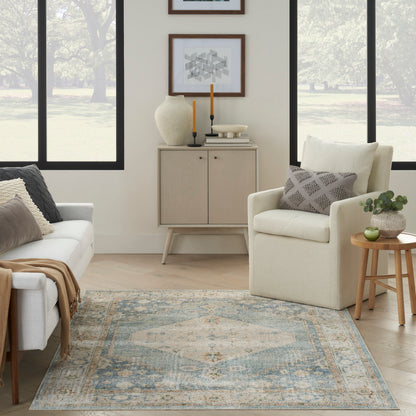 Neutral living room with beige armchair, light wood cabinet, blue distressed rug, round wood side table, potted plant, and framed geometric art.