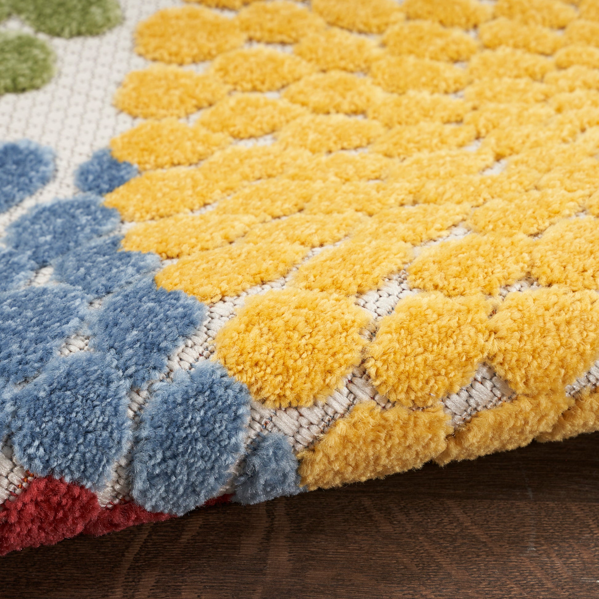Close-up of a plush tufted rug with yellow pom-pom clusters, blue accents and a red edge on a beige background.