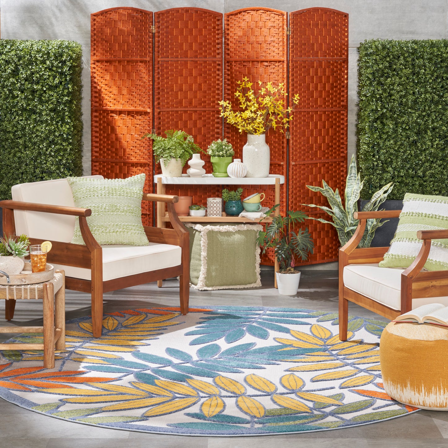Round tropical leaf rug in an outdoor seating area with wooden-frame chairs, beige cushions, green pillows, an orange folding screen and potted plants.