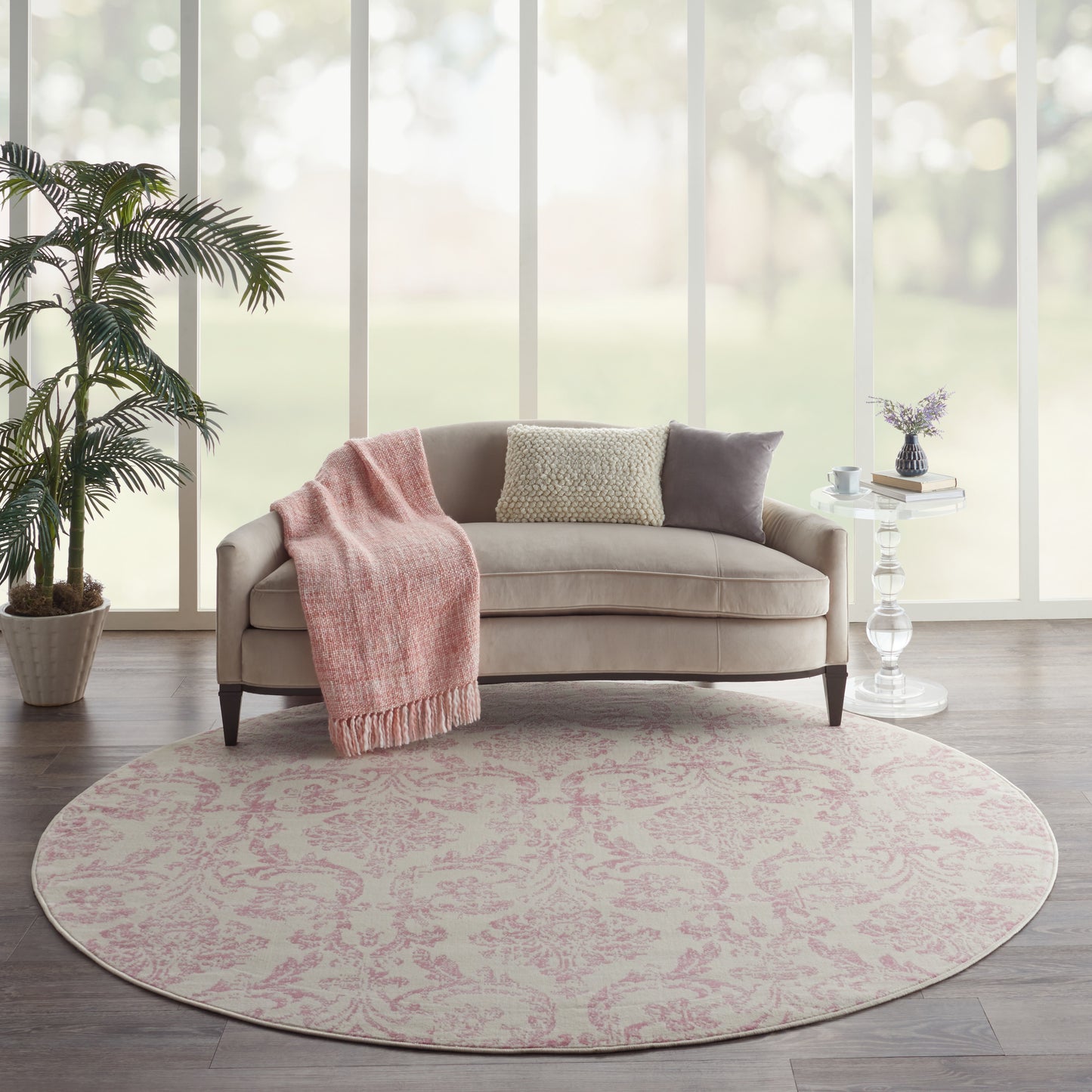 Round pink-patterned rug under beige sofa with blush throw, cushions, plant, and glass side table by large windows.
