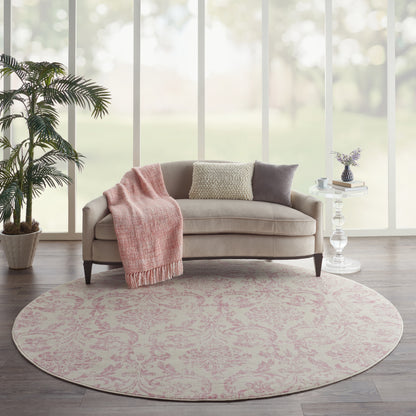 Round pink-patterned rug under beige sofa with blush throw, cushions, plant, and glass side table by large windows.
