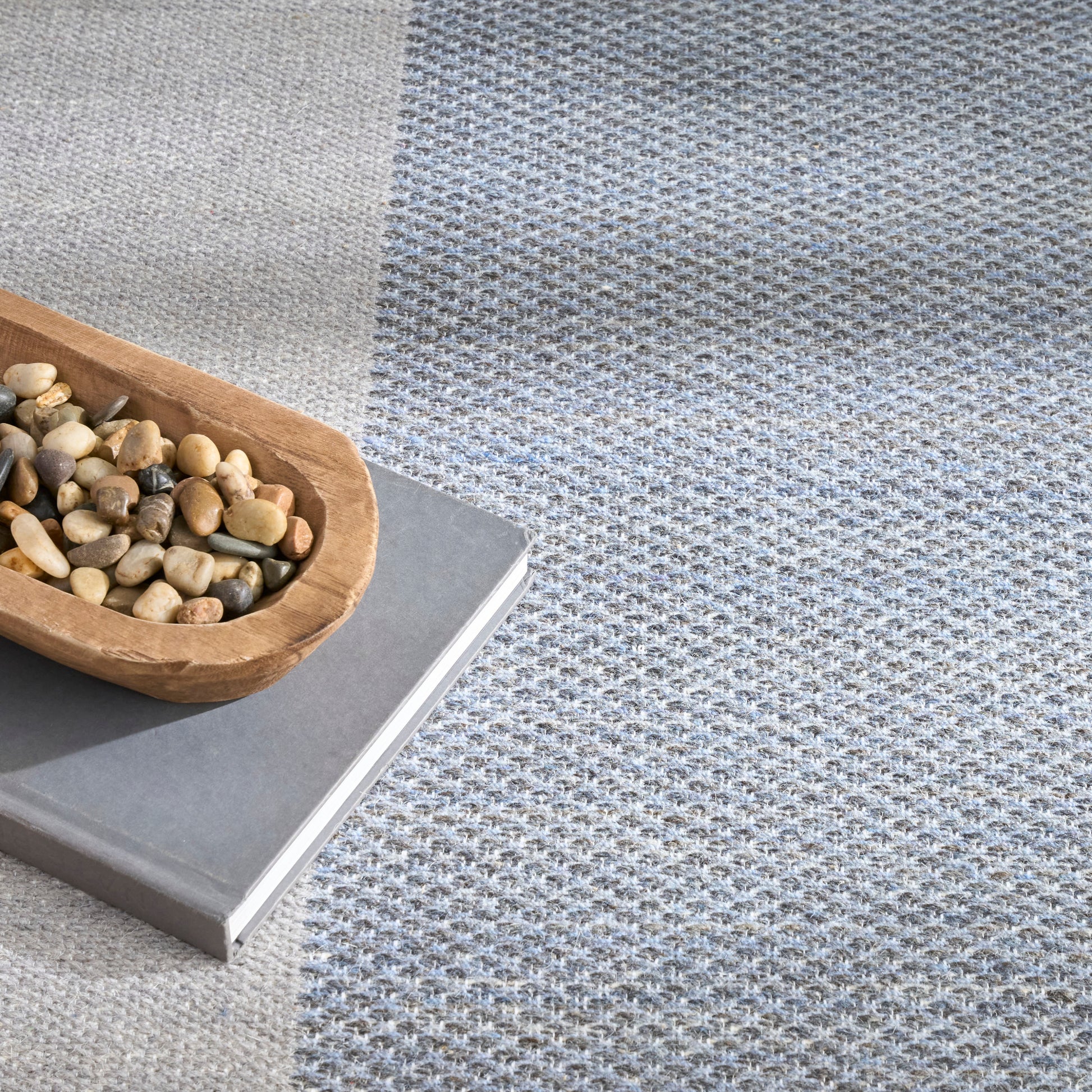 Two-tone woven rug in light gray and blue-gray with a wooden scoop of pebbles resting on a gray book.