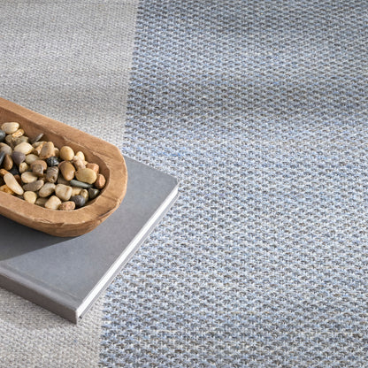 Two-tone woven rug in light gray and blue-gray with a wooden scoop of pebbles resting on a gray book.