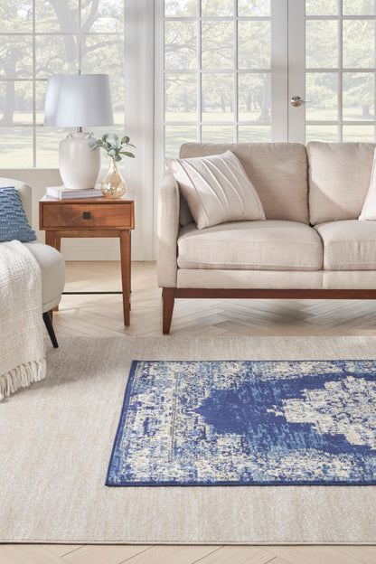 Beige sofa with cream cushions, wooden end table with lamp and vase, blue distressed rug on neutral beige floor.