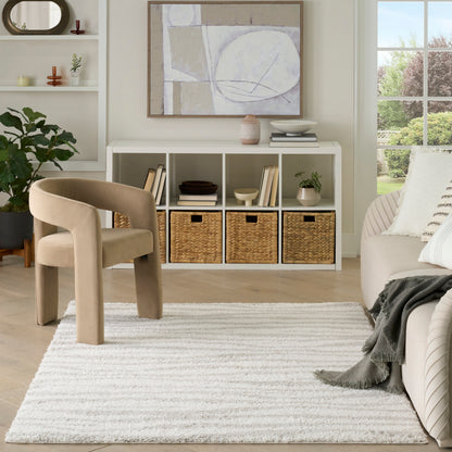 Neutral living room with beige curved chair, white cubby console with baskets, abstract art, plants, and a light shag rug.