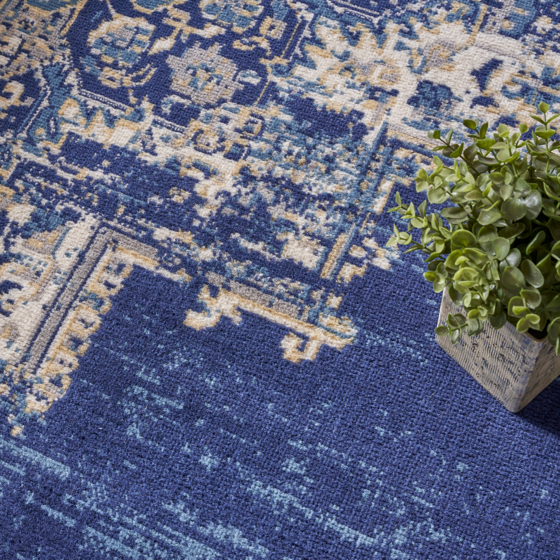 Blue distressed-pattern area rug with beige geometric motifs & woven texture; rectangular, shown with a small potted plant.