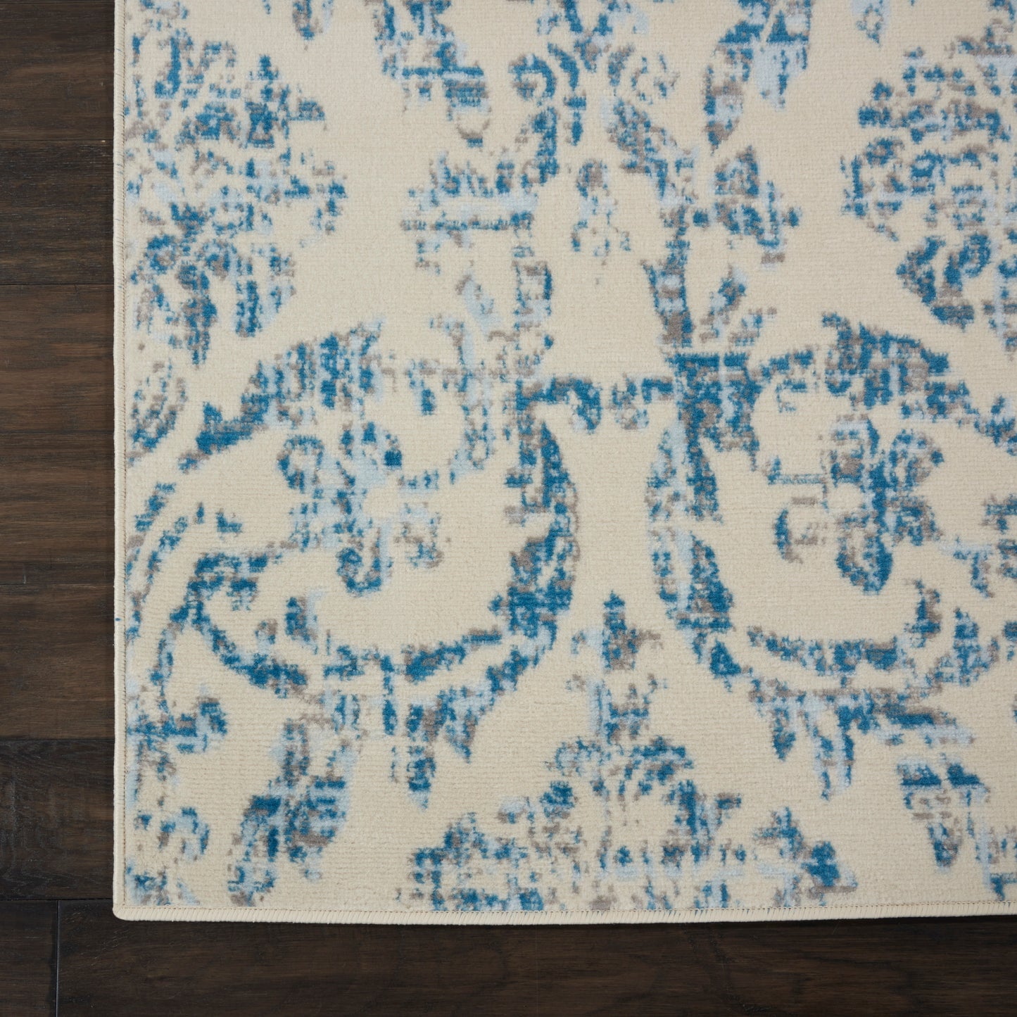 Distressed blue floral-pattern rug on light beige background, corner resting on dark wood floor.