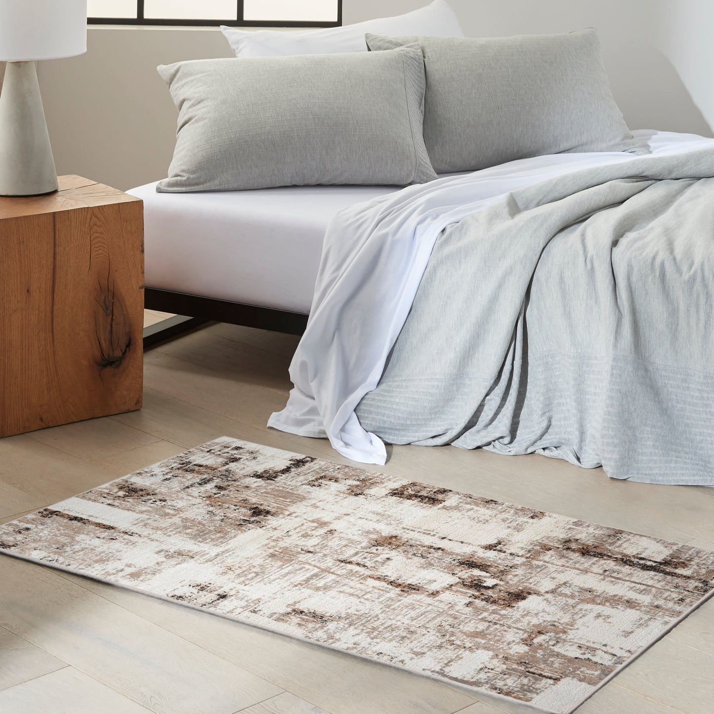 Cozy modern bedroom with light gray bedding and pillows, white sheets, a beige-brown rug, and a wooden bedside table.