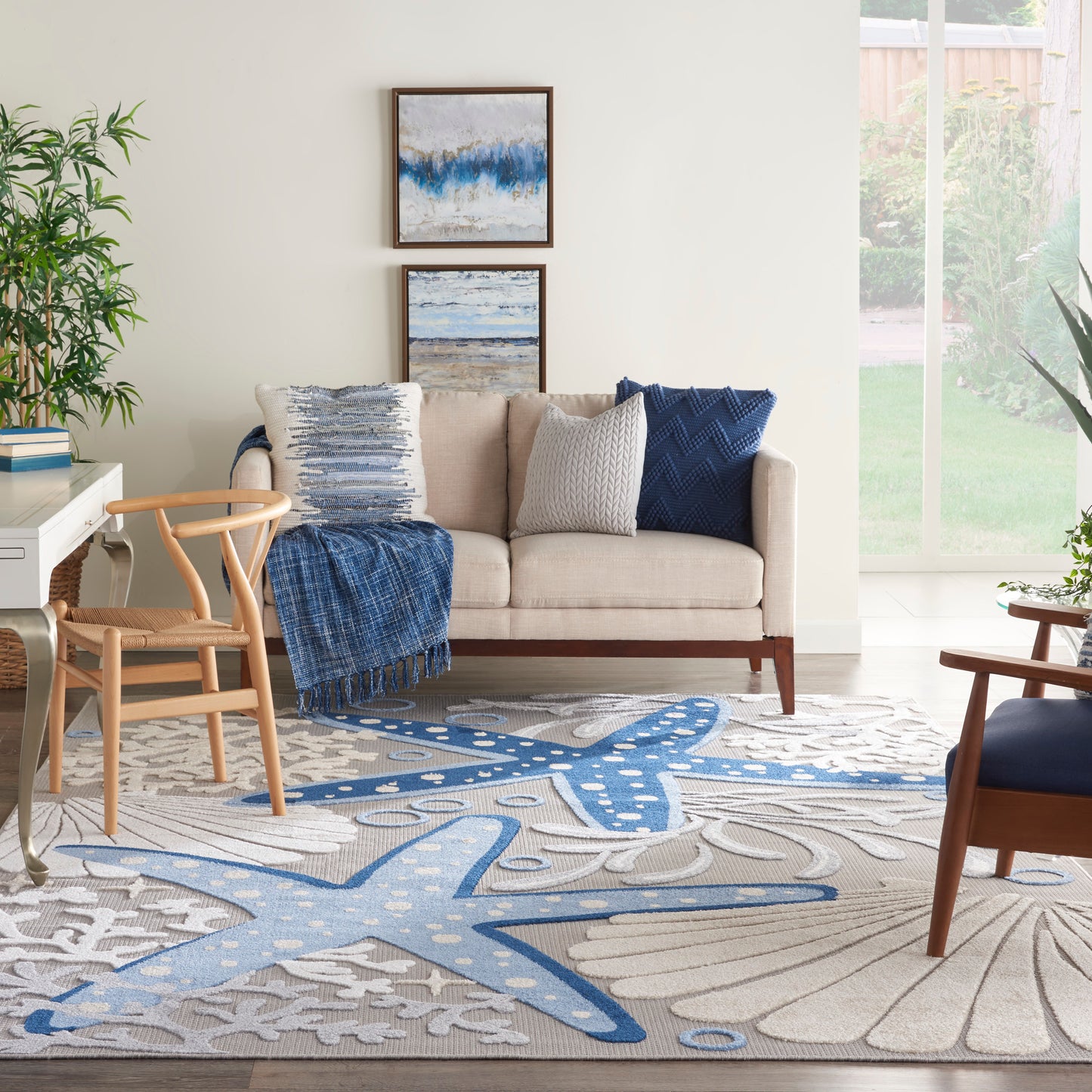 Coastal living room with beige sofa, blue throw, textured pillows, wooden chairs, framed art, and a blue-gray patterned rug.