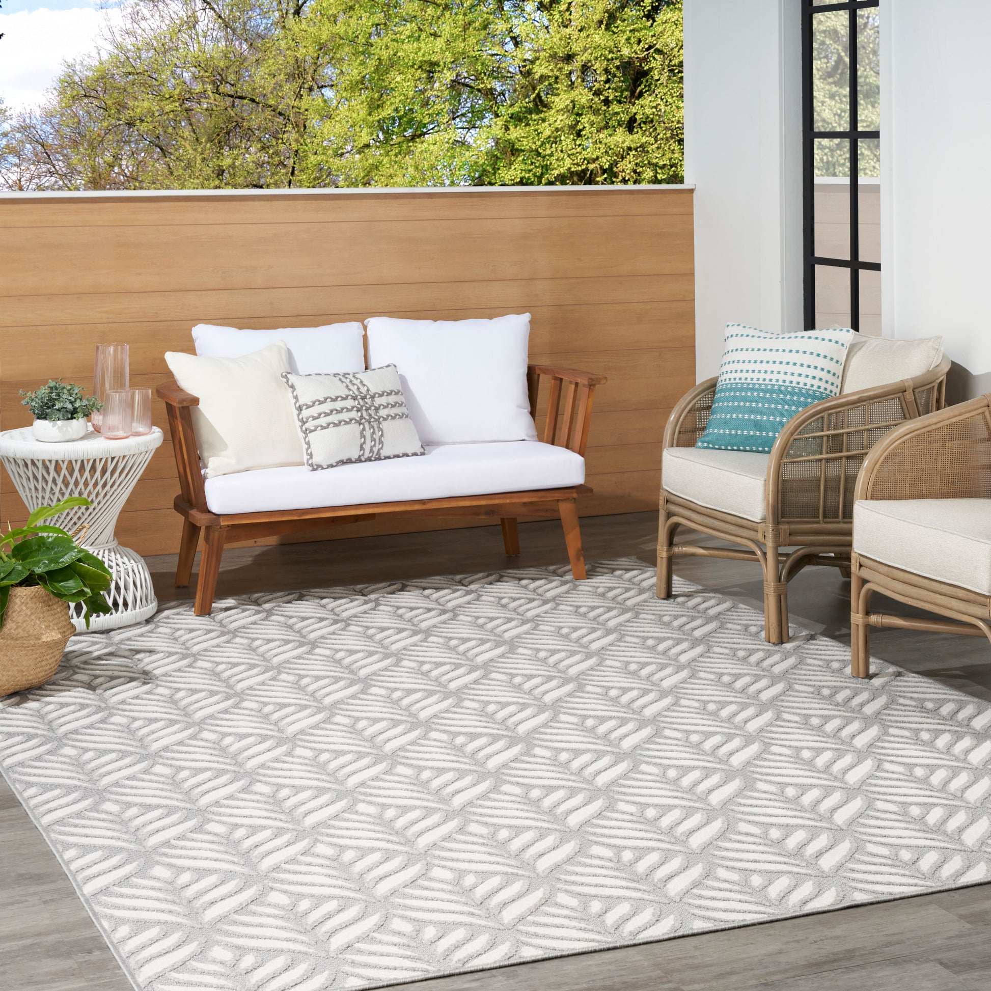 Outdoor patio with white cushioned wooden sofa, rattan chairs, side table, gray patterned rug, plants and privacy screen.