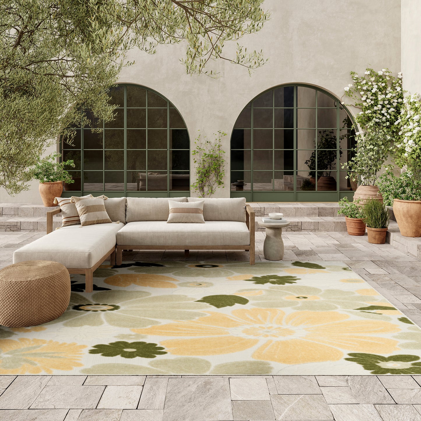 Neutral outdoor lounge with beige sectional, woven pouf, and a large yellow and green floral rug on a stone patio.