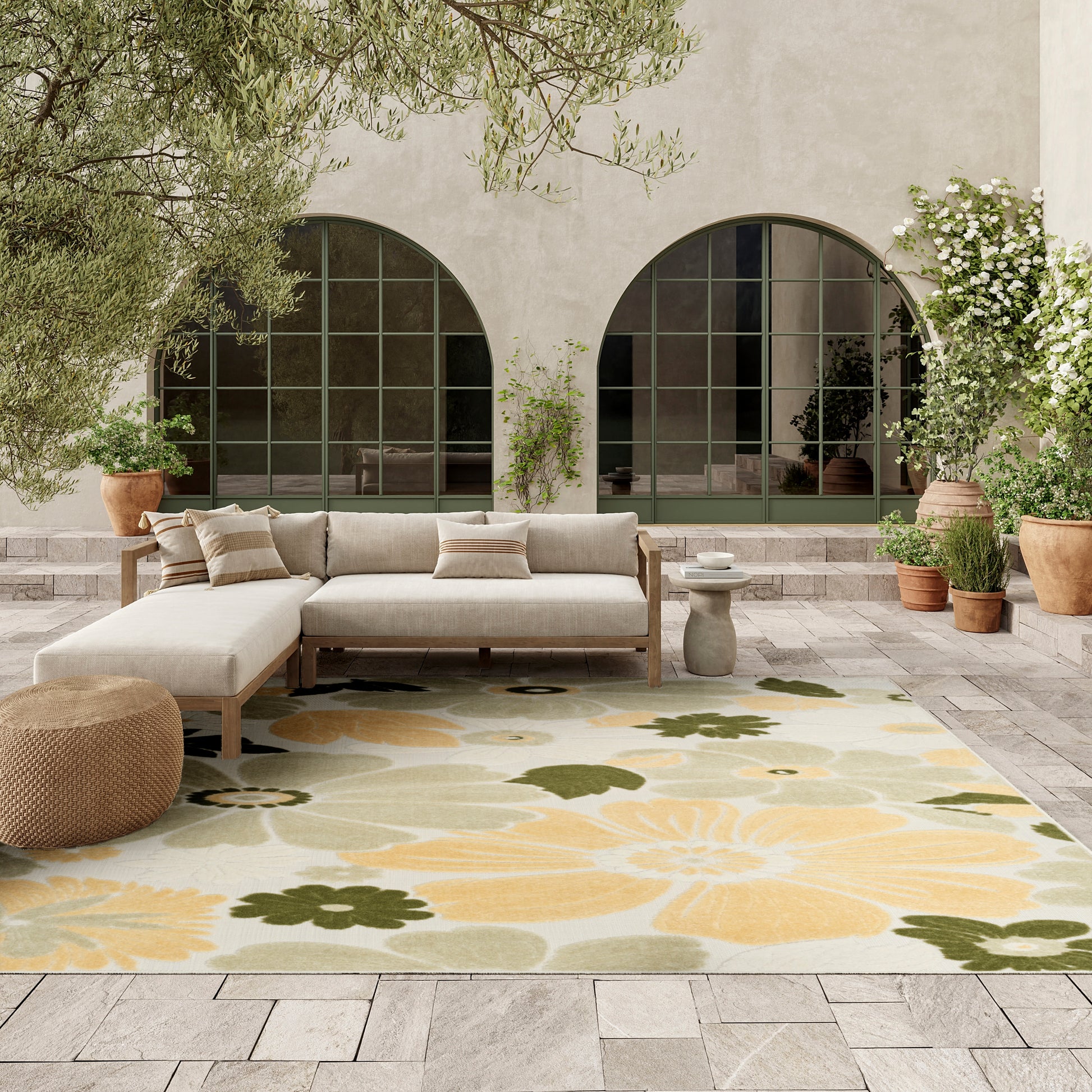 Neutral outdoor lounge with beige sectional, woven pouf, and a large yellow and green floral rug on a stone patio.