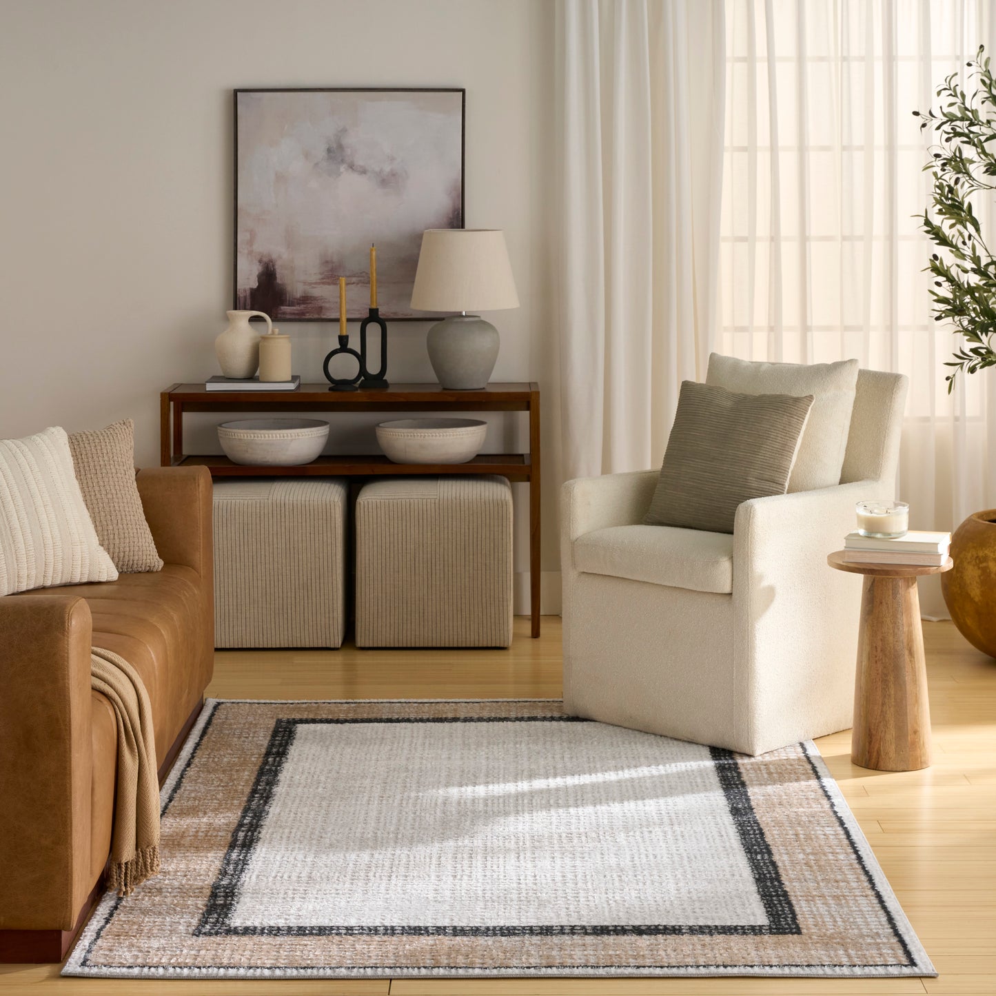 Neutral living room with an area rug, tan leather sofa, cream armchair, wooden console, beige ottomans, abstract art, and a ceramic lamp.