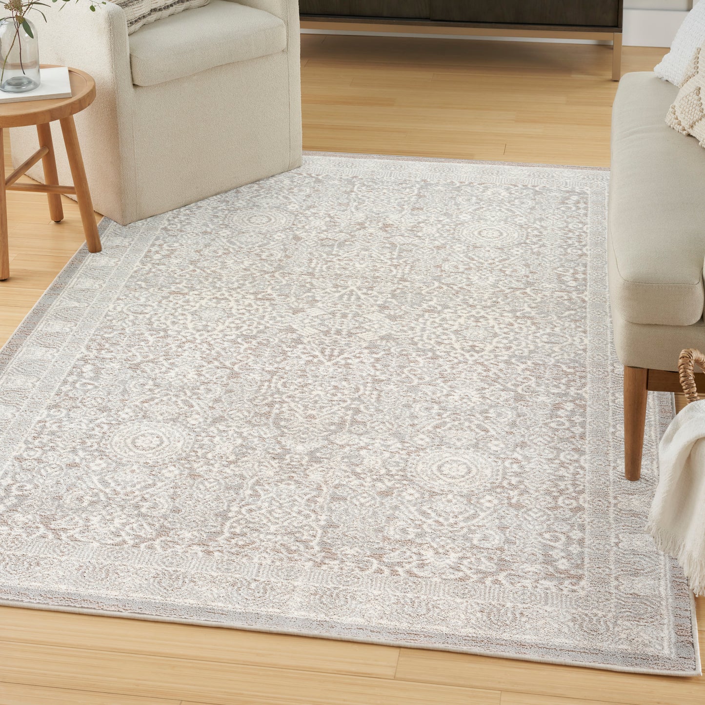 Beige ornate area rug with light gray pattern and border, laid over light wood flooring in a cozy living room.
